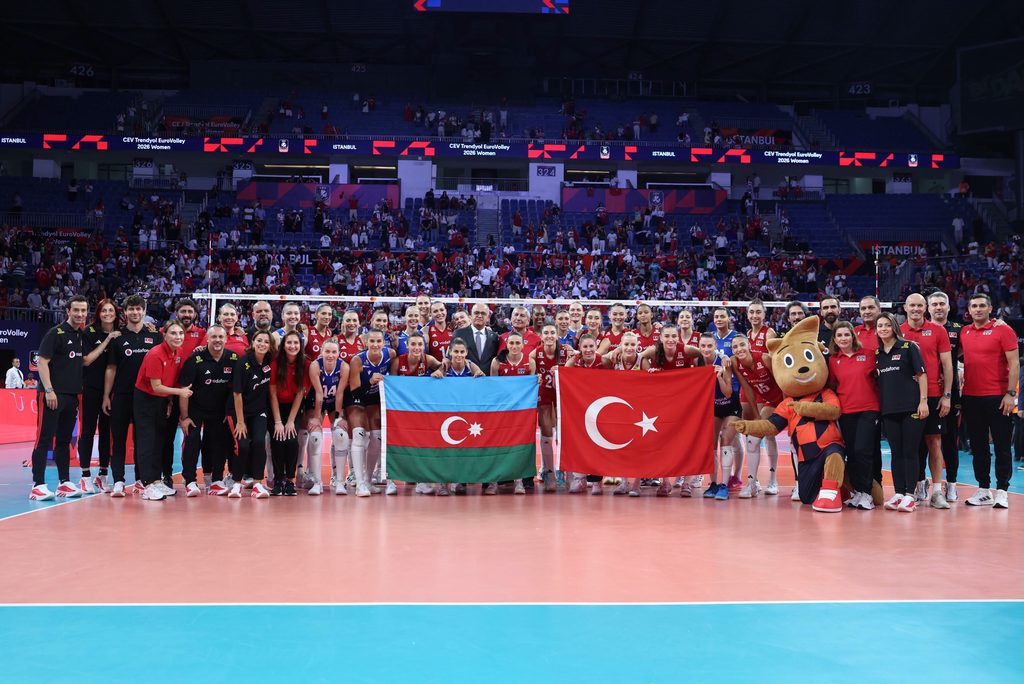 79167268823418112632541343791613213968546327n The Volley Lovers Group volleyball team posing for a photo with Turkish and Azerbaijani flags in a large indoor stadium with fans in the stands behind them.