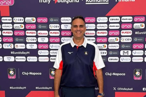 Man posing in front of a CEV EuroVolley 2026 backdrop, wearing a navy polo with white sleeves and dark pants.
