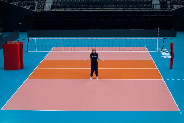 81397608810605462901071368601725172856851211n The Volley Lovers Person standing with arms outstretched on a colorful indoor volleyball court in front of a net, empty stands in the background