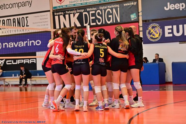 A group of female volleyball players in red and black uniforms huddle together on an indoor court, with banners and spectators in the background.