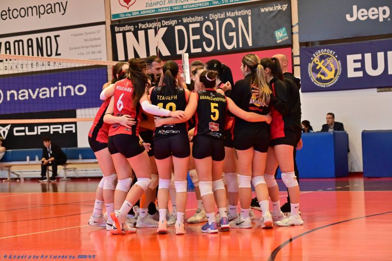 amazoneswn1 The Volley Lovers A group of female volleyball players in red and black uniforms huddle together on an indoor court, with banners and spectators in the background.