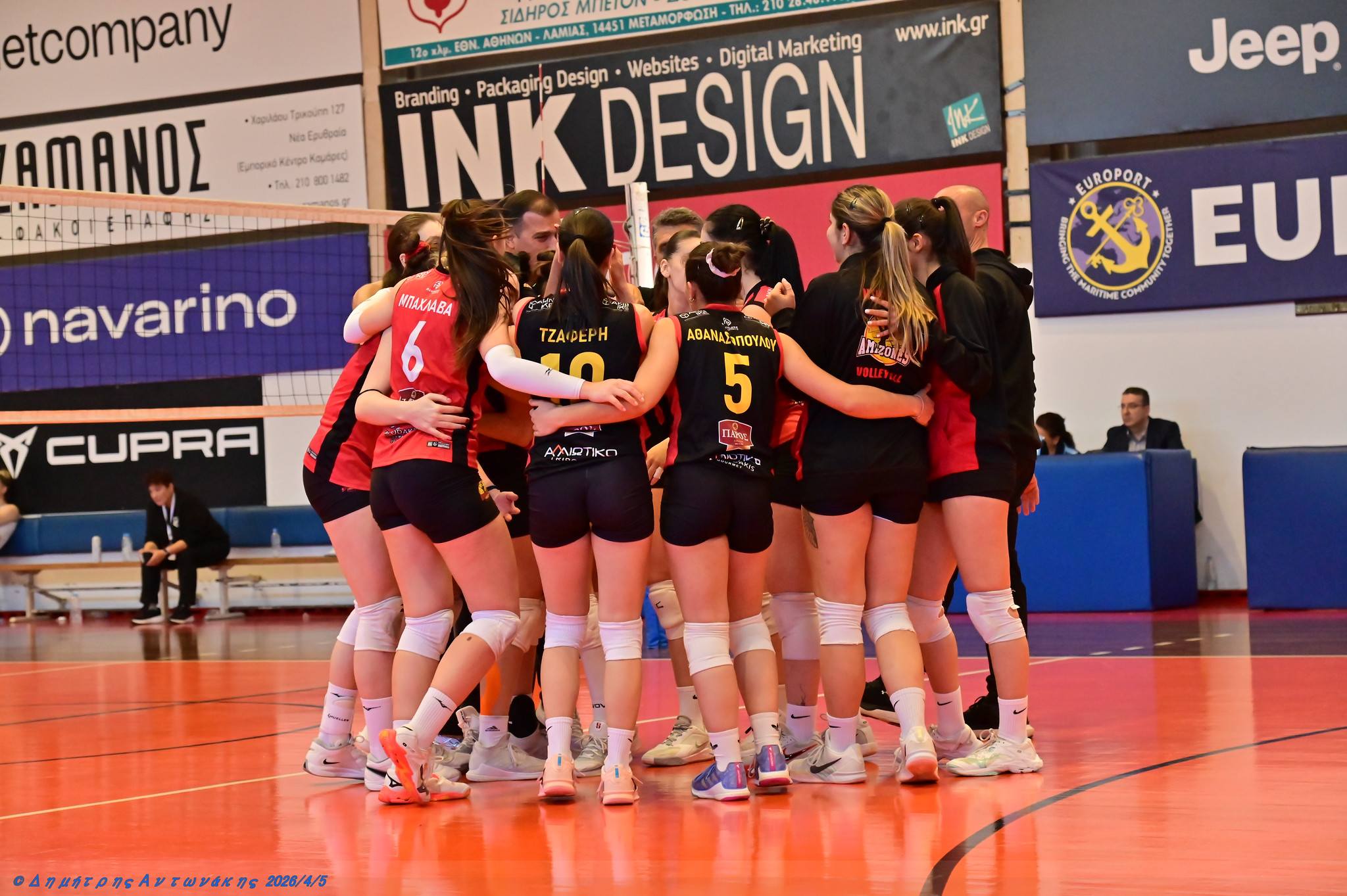 amazoneswn1 The Volley Lovers A group of female volleyball players in red and black uniforms huddle together on an indoor court, with banners and spectators in the background.