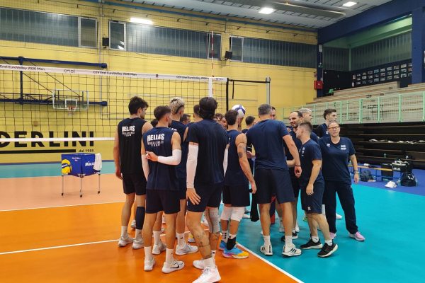 Indoor volleyball team huddle on a colorful court, players in blue uniforms forming a circle with coach nearby.
