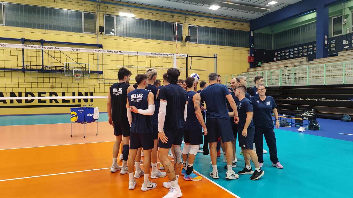 Indoor volleyball team huddle on a colorful court, players in blue uniforms forming a circle with coach nearby.