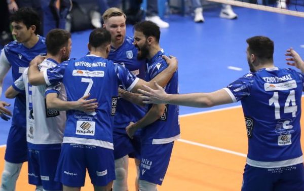 Blue volleyball players in a huddle on an orange court, celebrating with arms around each other during a match.