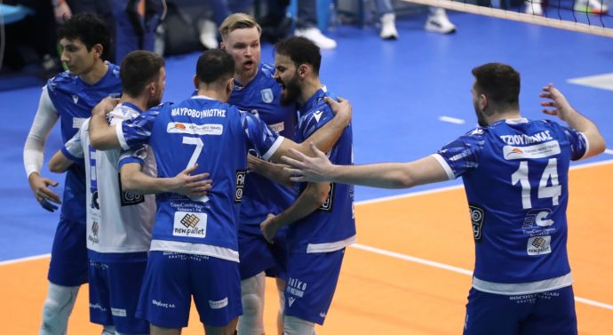 iramen The Volley Lovers Blue volleyball players in a huddle on an orange court, celebrating with arms around each other during a match.