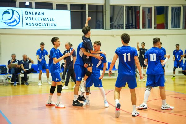 Teenage volleyball players in blue uniforms celebrate on an indoor court, one lifting a teammate as others cheer; Balkan Volleyball Association banner visible in the background.