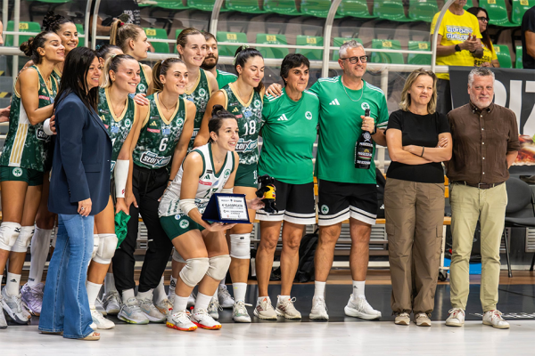 A women's volleyball team poses with a trophy and coach staff after a match in an indoor arena.