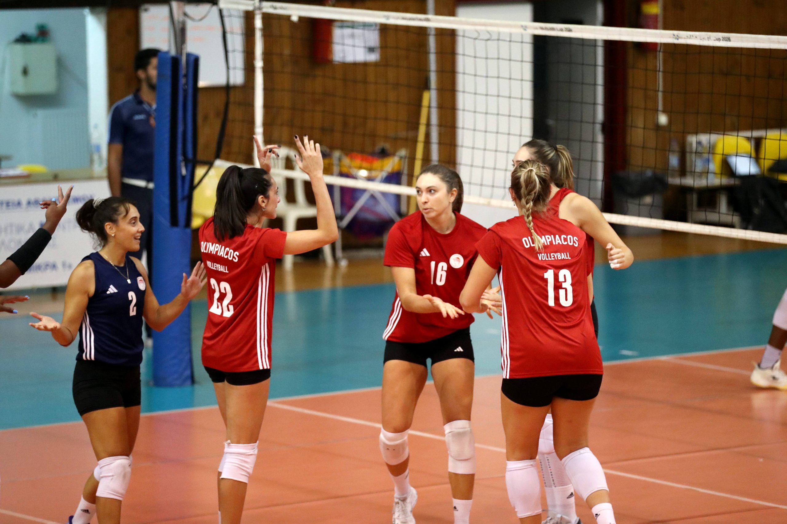 Group of female volleyball players in red jerseys celebrate on an indoor court near the net.