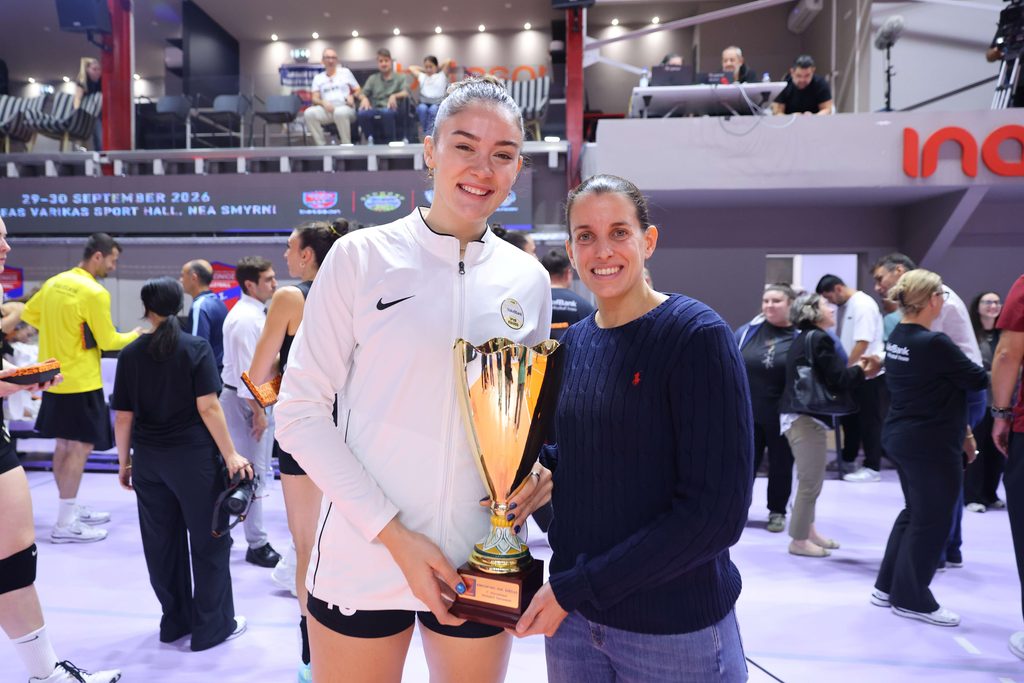 005359842 The Volley Lovers Two smiling women pose with a large trophy at an indoor sports event, with a crowd and banners in the background.