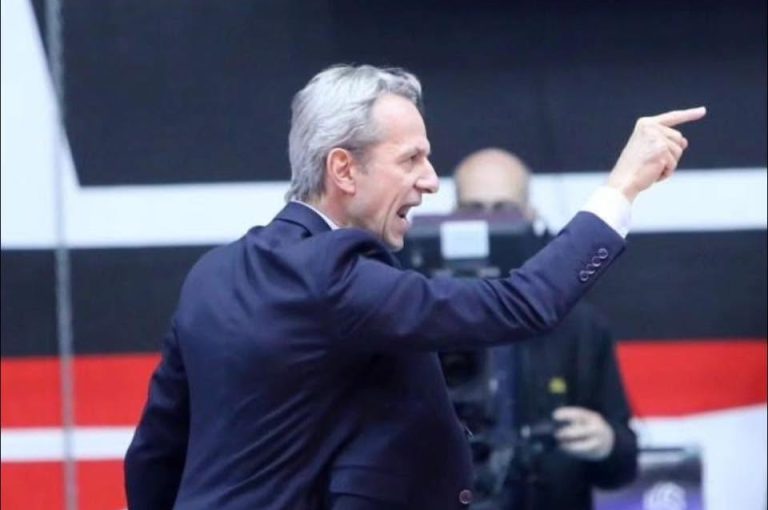 Man in a dark suit on stage, gesturing with his raised right arm and pointed finger toward the audience. (Informative image showing a speaker during an event)