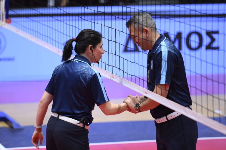 Intime Kyriazis Stefanos The Volley Lovers Two officials or players in navy uniforms shake hands at a volleyball net inside a gym.