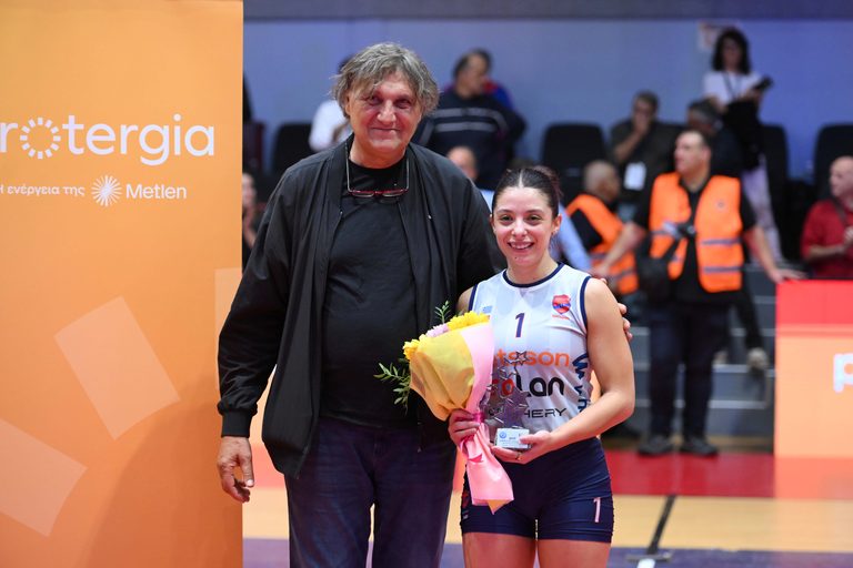 537815400 The Volley Lovers Older man in a black jacket standing beside a smiling female athlete in a white-and-blue jersey holding a bouquet at a sports event.