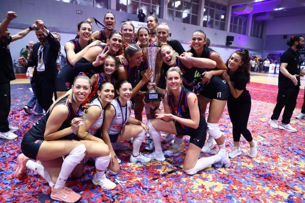 Female basketball team celebrates victory, posing with a large trophy and medals on a confetti-covered court.