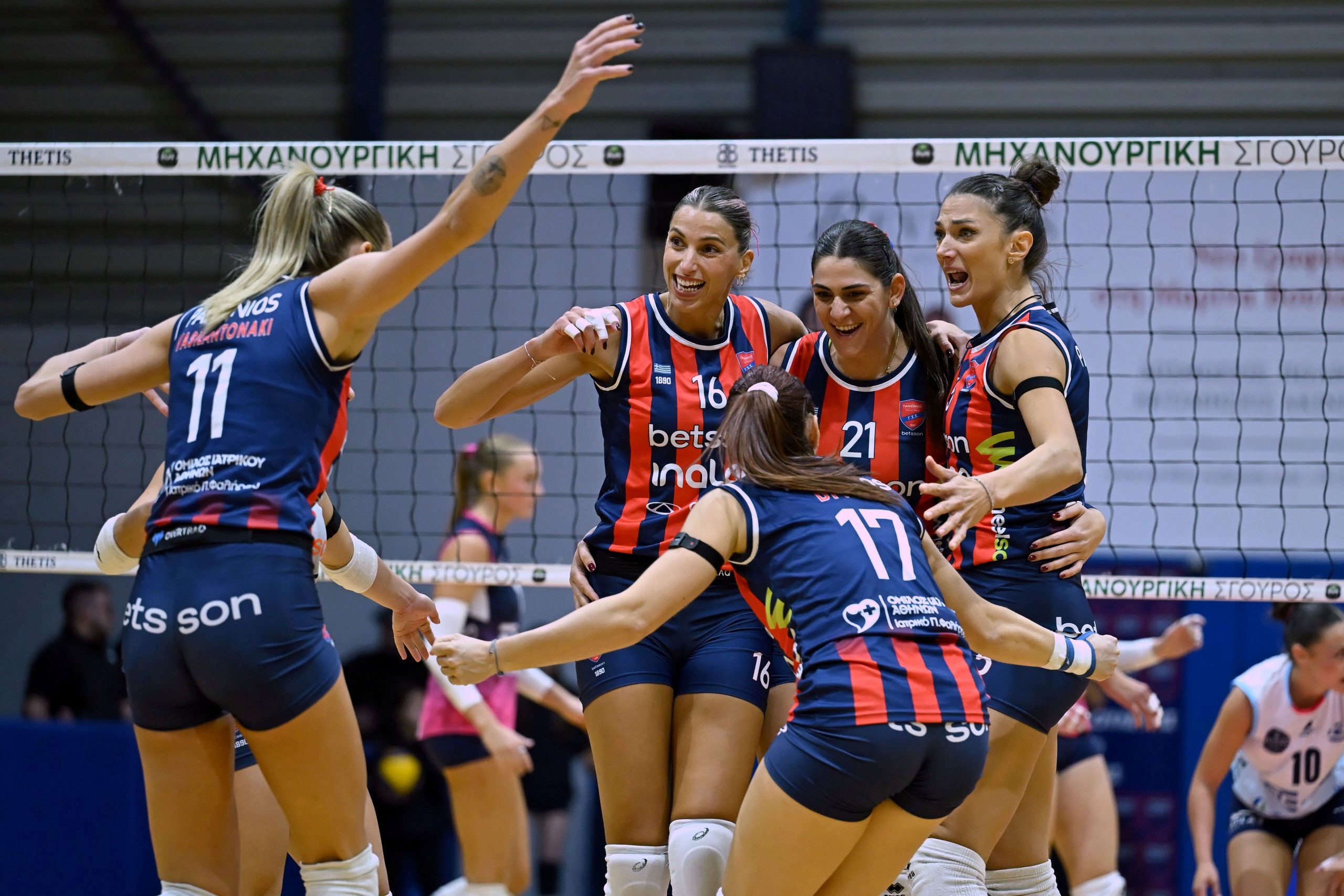 538982700 The Volley Lovers A group of female volleyball players in blue-and-red uniforms celebrate at the net, arms raised in joy after a point.