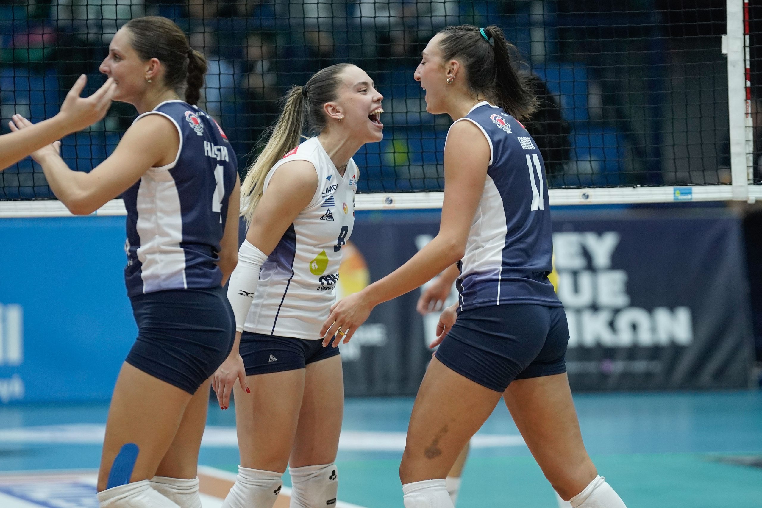 Intime Karanikolos Harris The Volley Lovers Three female volleyball players in navy and white celebrate a point near the net on an indoor court.
