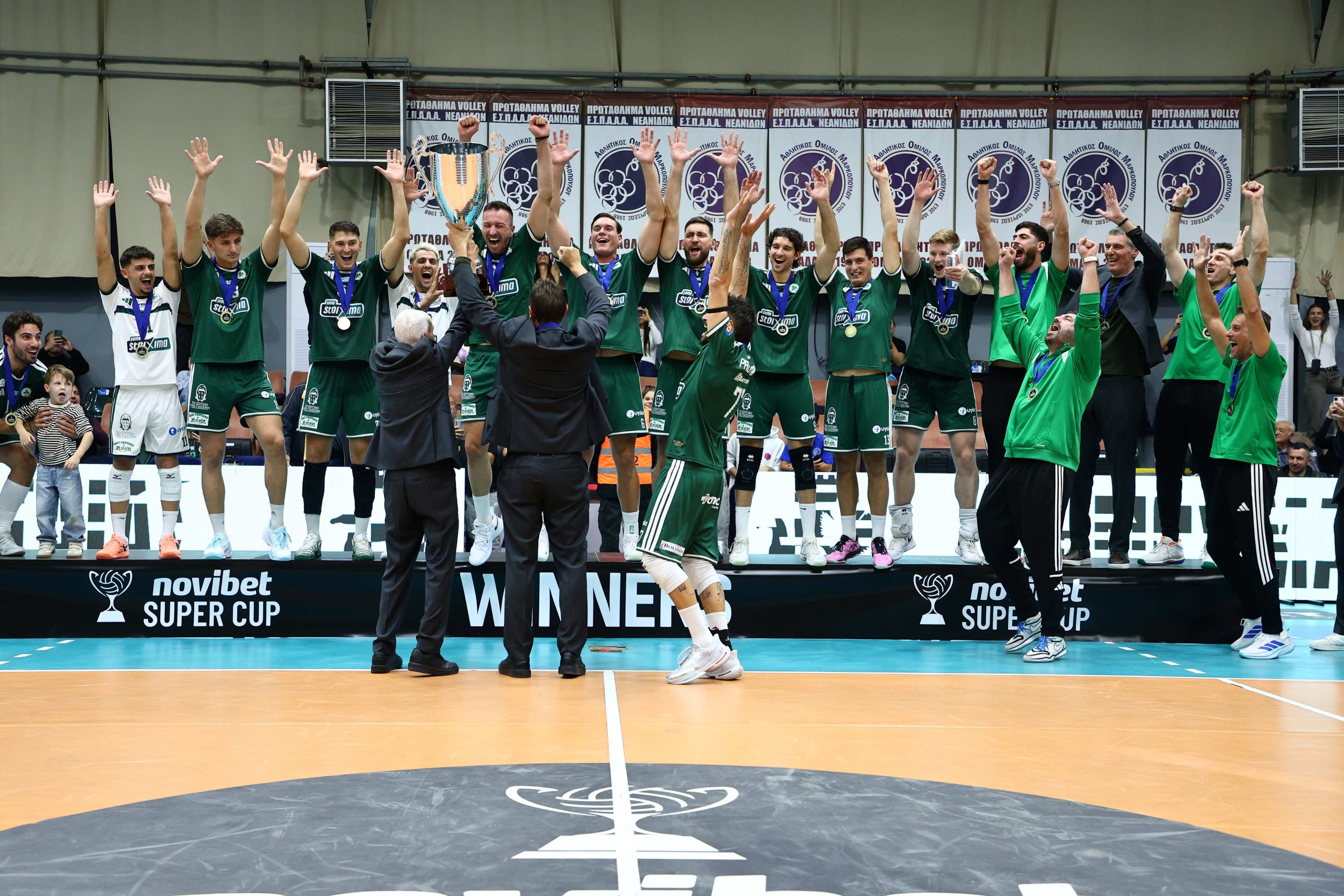 A volleyball team in green uniforms raises arms in victory around a large trophy on the podium. Medals hang from their necks as staff applaud in the foreground.