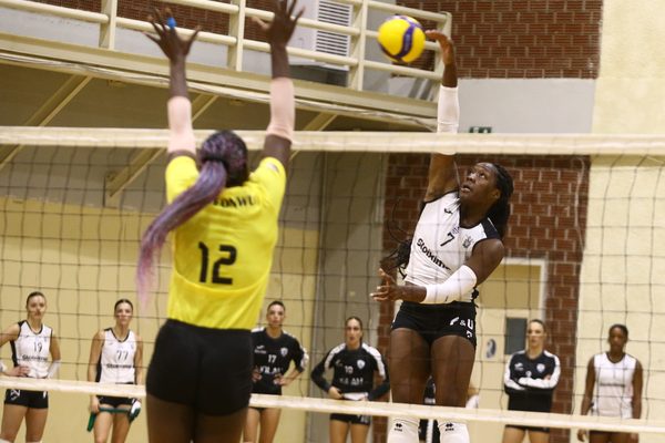 Right-side player in white jersey leaps to spike the ball over the net while a yellow-jersey defender jumps to block it.