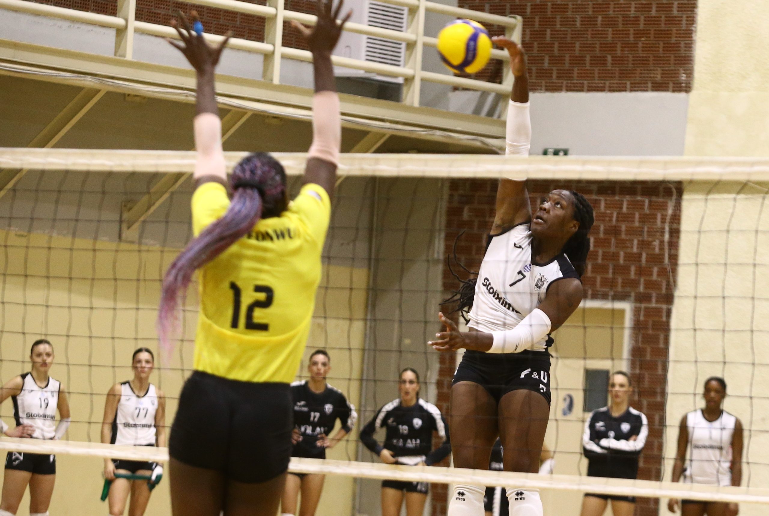 Right-side player in white jersey leaps to spike the ball over the net while a yellow-jersey defender jumps to block it.