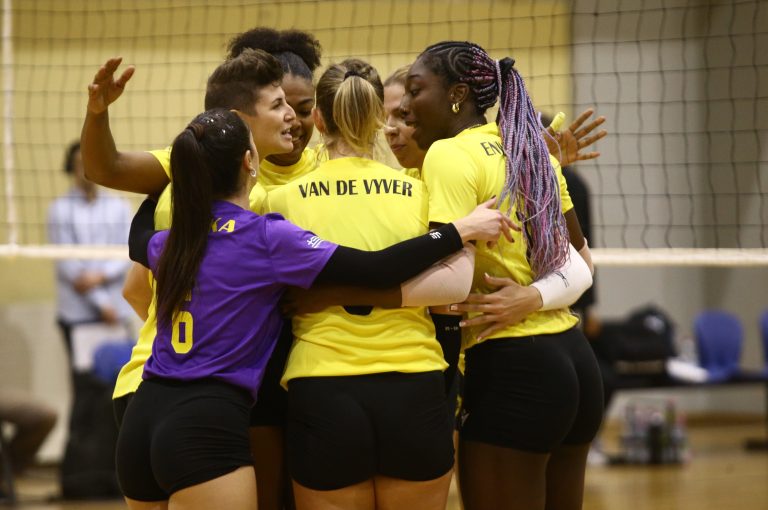 A group of volleyball players in yellow jerseys huddles together in celebration on an indoor court, with one player in purple joining the embrace.