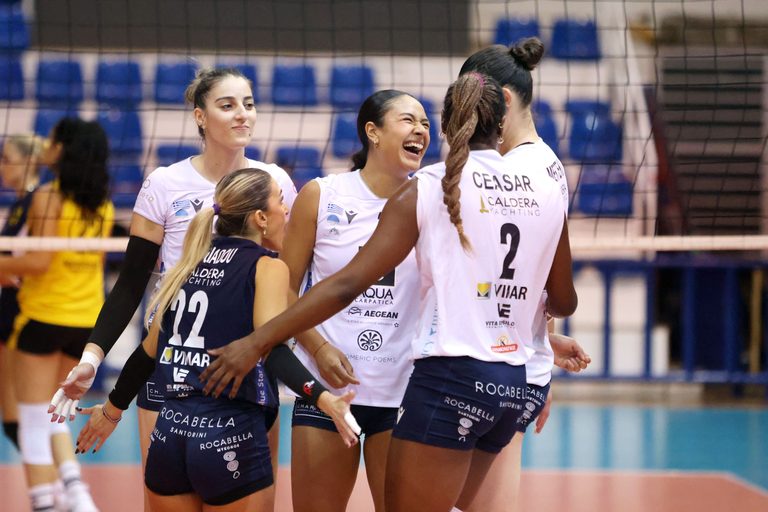 Group of female volleyball players in white uniforms smiling and embracing on the court beside a net.