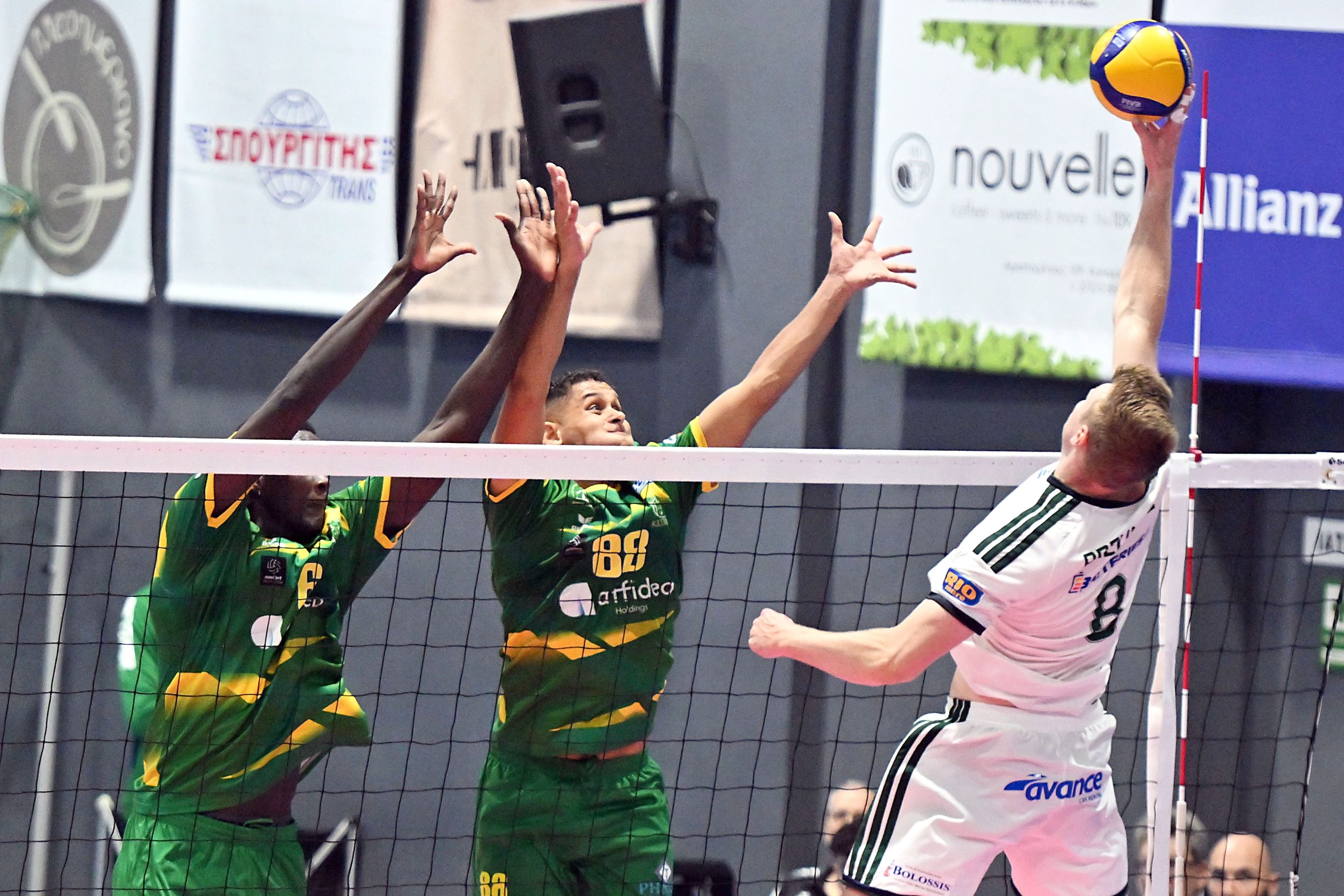 Volleyball players in green jump at the net to block as a white-jersey opponent attacks; yellow ball overhead.