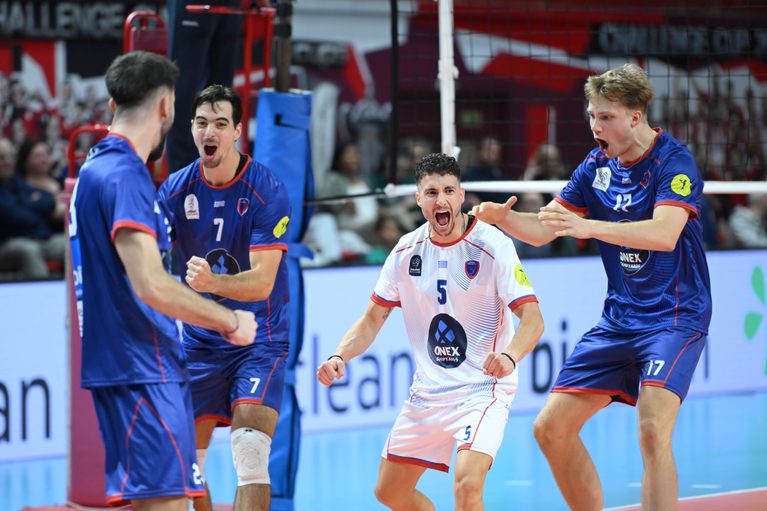 Volleyball players in blue uniforms celebrate a point near the net, center player in a white jersey shouting with fists raised.