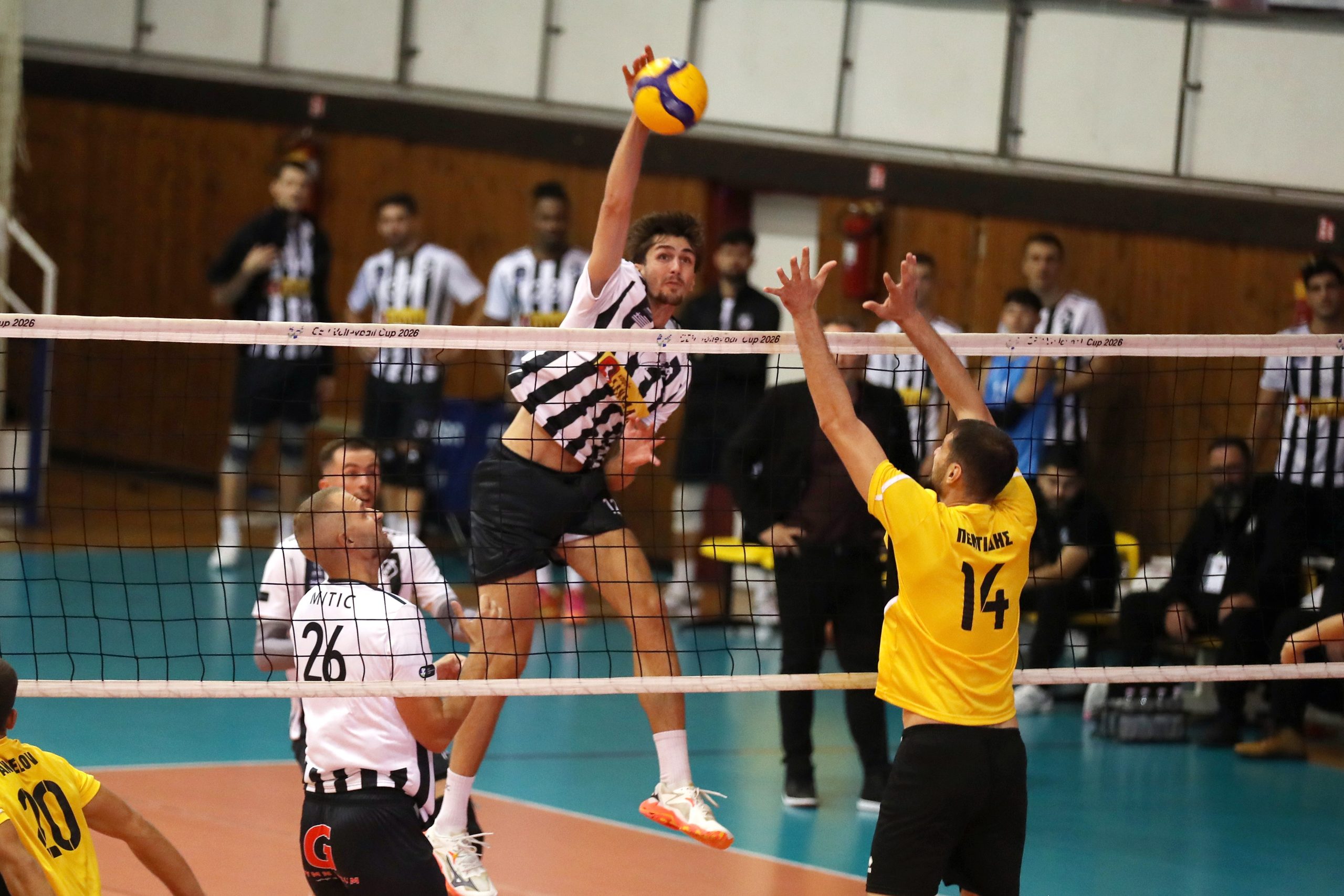 Male volleyball player in a black-and-white striped jersey jumps to spike a yellow volleyball over the net as a defender in a yellow jersey reaches up in front of him with spectators in the background