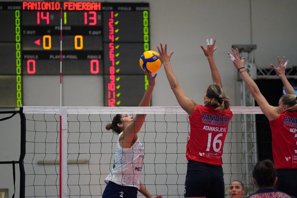 55555584413033646315775042620379440155116812n The Volley Lovers Volleyball rally: red-jersey players jump to block at the net as a white-clad player hits the ball; scoreboard in the background.