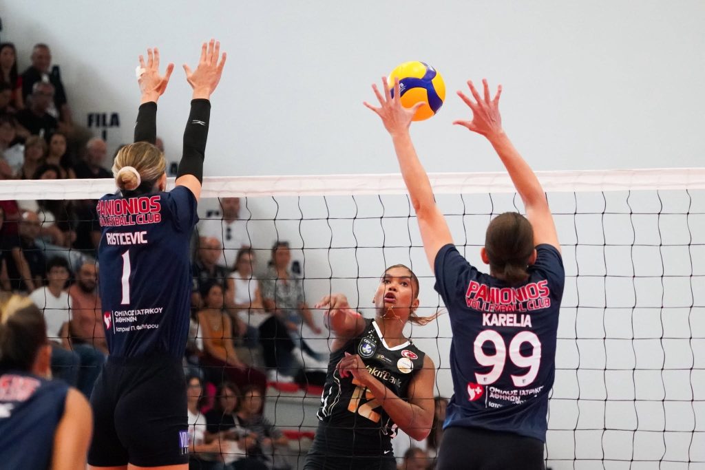 55597152713039090748563937404387164796236696n The Volley Lovers Volleyball players jump to block a ball at the net while a crowd watches from the stands.