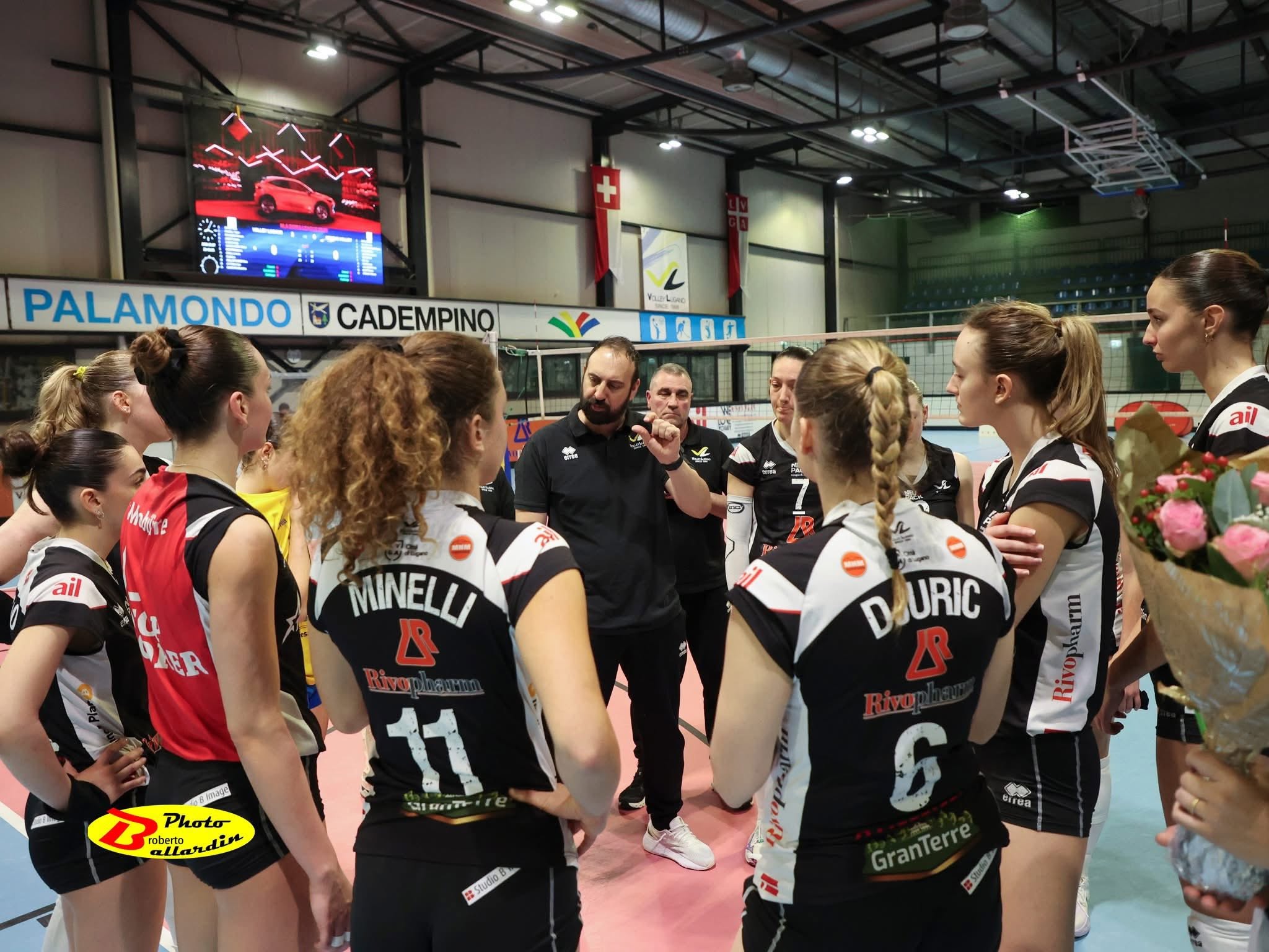 Female volleyball players form a huddle in a gym, wearing black-and-white uniforms with numbers, listening to coaches. Background shows banners and a scoreboard.