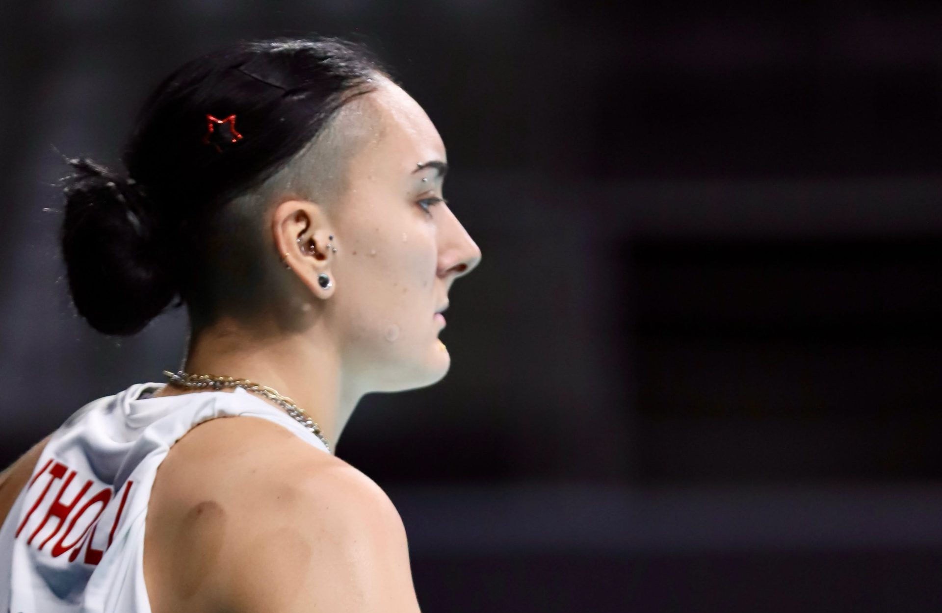 Side profile of a person with short, wet hair tied back, wearing a white sleeveless top, and visible water droplets on the face and neck, looking to the right.