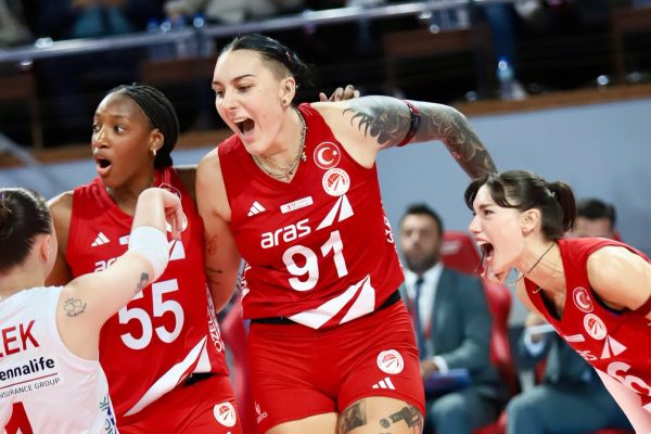 Female volleyball players in red uniforms celebrate together on court, arms raised in victory.