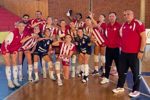 Women’s volleyball team posing together after a match, many in red-and-white striped uniforms in a gym. Coaches stand at right.
