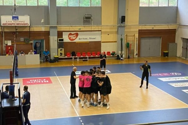 Volleyball team in pink and black jerseys huddles on an indoor court near the net, a coach guiding them. A referee and bench area are nearby.