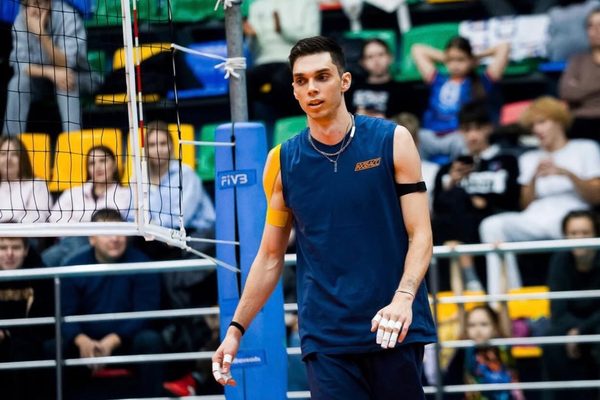 Male indoor volleyball player in a blue sleeveless jersey stands near the net with a cheering audience in the stands behind him.