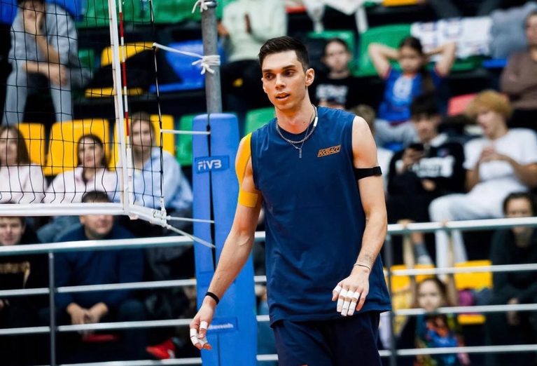 Male indoor volleyball player in a blue sleeveless jersey stands near the net with a cheering audience in the stands behind him.