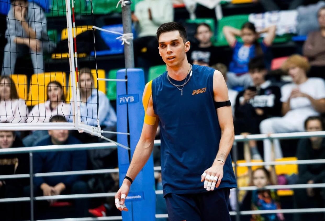 Male indoor volleyball player in a blue sleeveless jersey stands near the net with a cheering audience in the stands behind him.