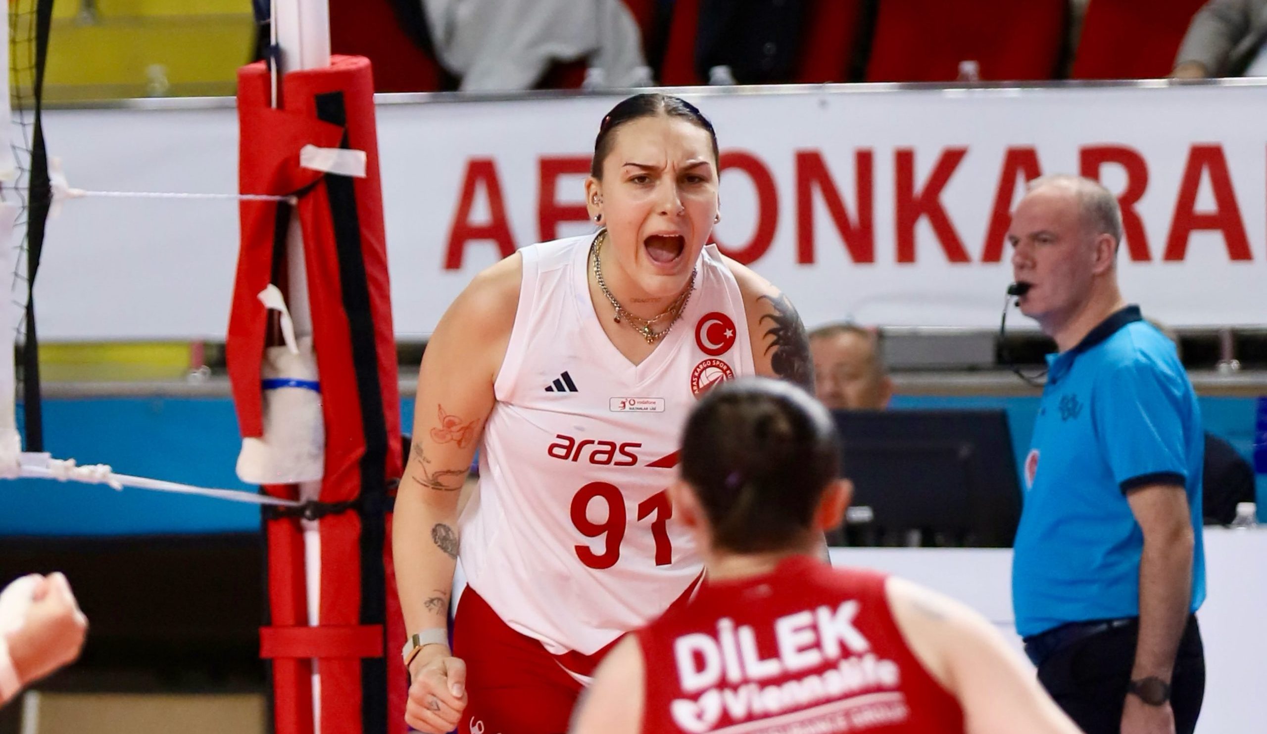 Female volleyball player in white and red jersey screams during a rally near the net, with red padding and spectators behind.
