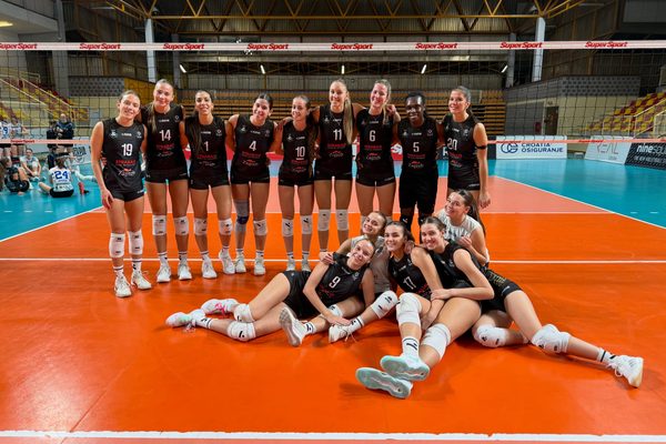 Group of female volleyball players in black uniforms posing together on an indoor court with a net behind them.
