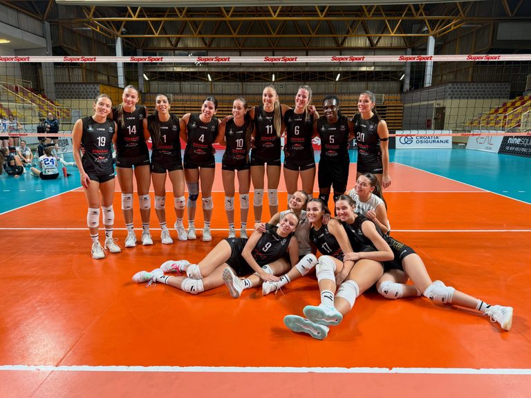856854612184320544961974841609197779711198467n The Volley Lovers Group of female volleyball players in black uniforms posing together on an indoor court with a net behind them.
