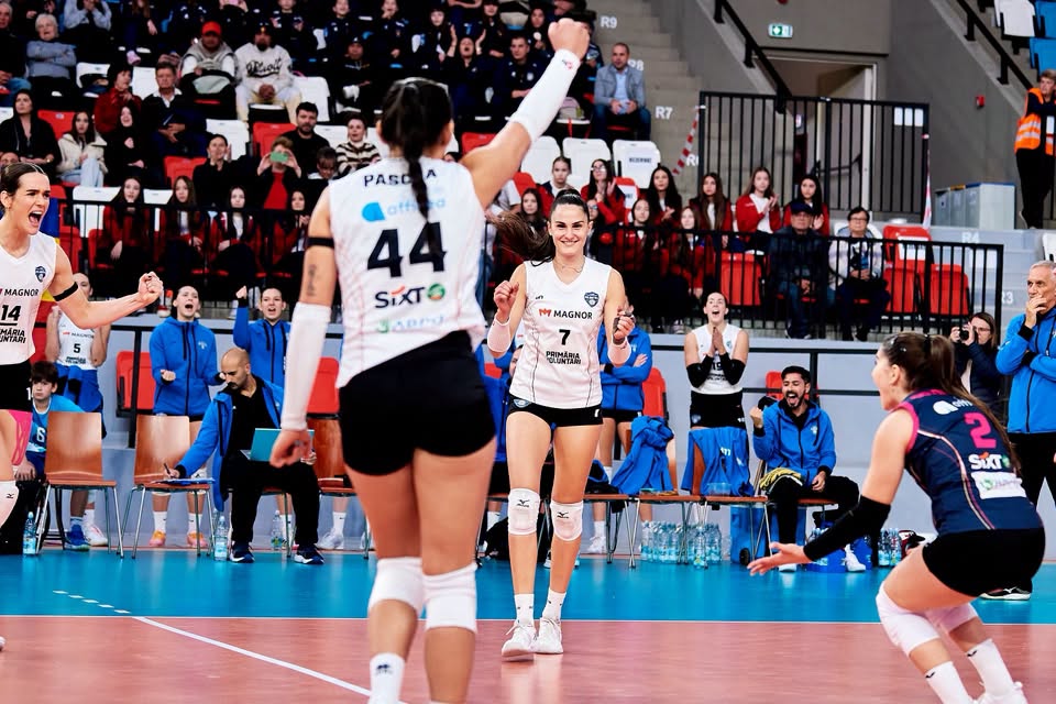 Indoor volleyball match scene with players celebrating a point as fans cheer in the stands behind them, one player facing camera with a smile and others raising fists in triumph
