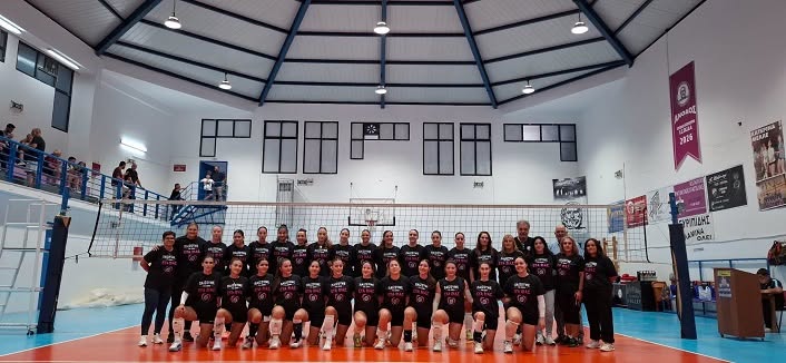 Women’s volleyball team posing for a group photo on an indoor court with a net and banners in the gym stands behind them, everyone wearing matching black outfits.