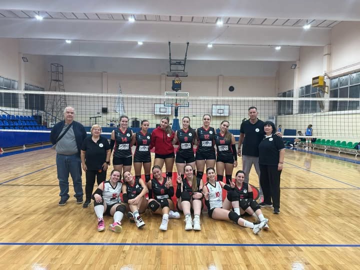 Girls' volleyball team and two coaches pose for a group photo on an indoor court with a net, wearing black and red uniforms.