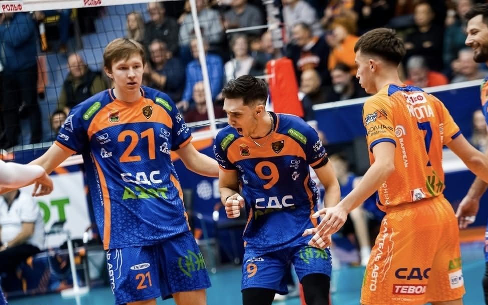 Volleyball players in blue and orange jerseys celebrate on the court as spectators watch from the stands.