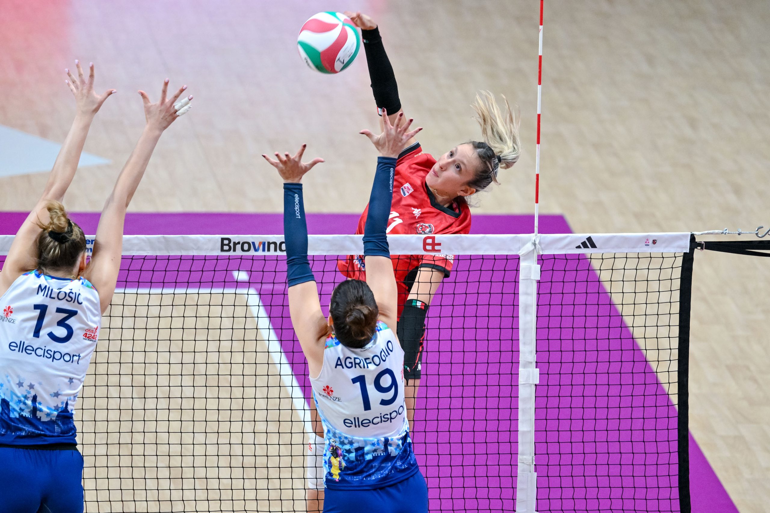 Female volleyball players at the net: one in red spikes the ball as others jump to block, during a match.