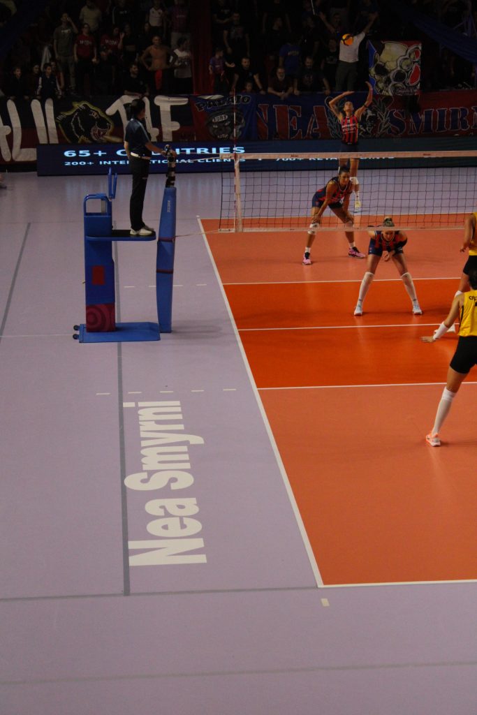 IMG3088 The Volley Lovers Volleyball match on an orange court with a referee on a raised platform and players near the net, spectators in the stands behind a bannered wall.