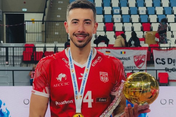 Smiling athlete in a red jersey holds a trophy and medal in a confetti-strewn arena, stadium seating in background.