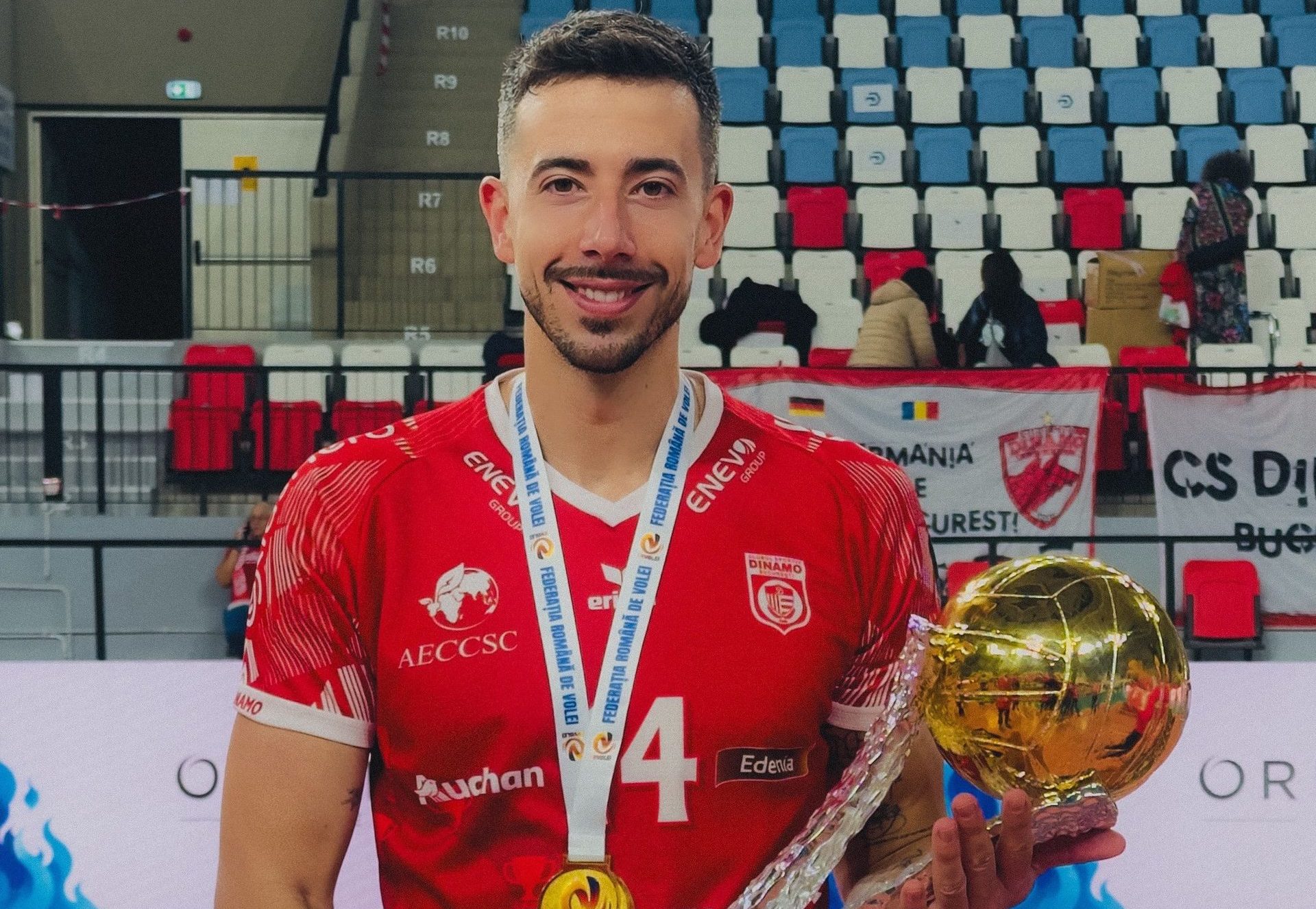 Smiling athlete in a red jersey holds a trophy and medal in a confetti-strewn arena, stadium seating in background.