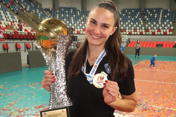 Smiling woman in a black shirt holds a tall silver trophy with a gold sphere on top and wears a round medal around her neck, confetti on the floor behind her in an arena.